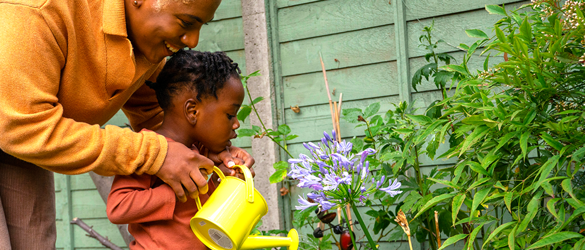 adult and child using a bright yellow watering can to water a plant outside