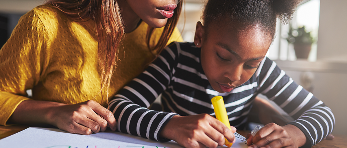 Young girl writing in coloured pens on a large sheet of paper. An adult woman is sitting behind the girl leaning over her shoulder and looking at what the girl is writing.