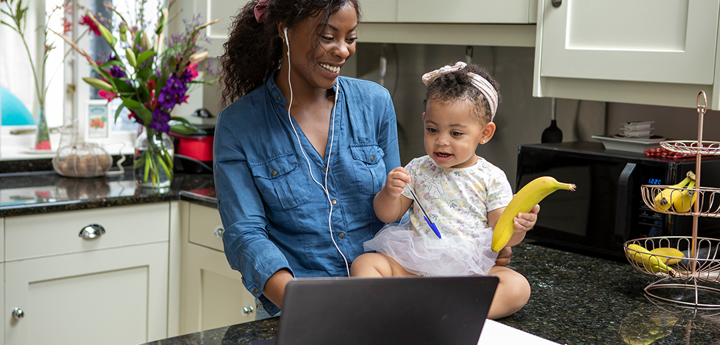 Toddler holding a banana sitting on kitchen countertop supported by parent. Parent has earphones in and both are in front of a laptop.