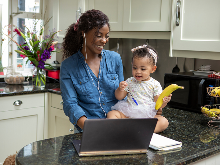 Toddler holding a banana sitting on kitchen countertop supported by parent. Parent has earphones in and both are in front of a laptop.