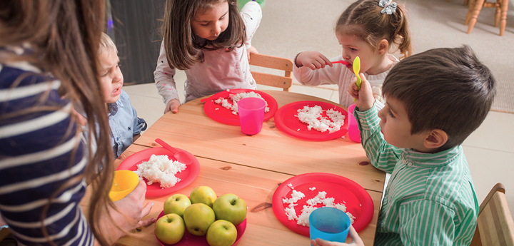 Group of 4 young children sitting round a table eating food from plastic plates.