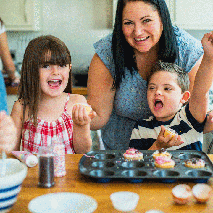 2 children and a woman making cupcakes at a kitchen bench. The boy is throwing his fist into the air excitedly whilst the girl is grinning.