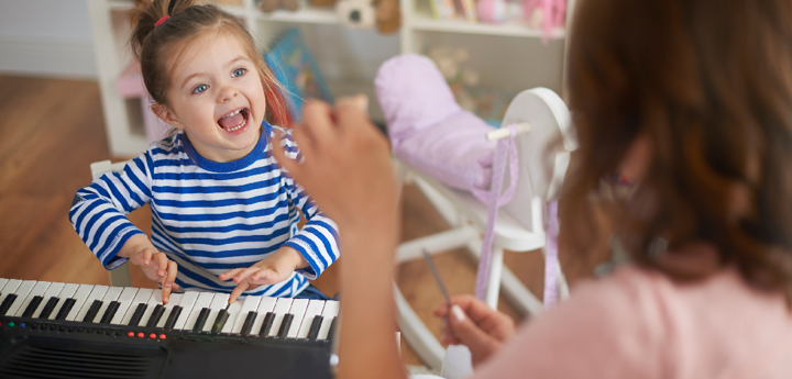 Young girl smiling whilst pushing keys on a keyboard. A woman in the foreground is facing the girl and conducting.