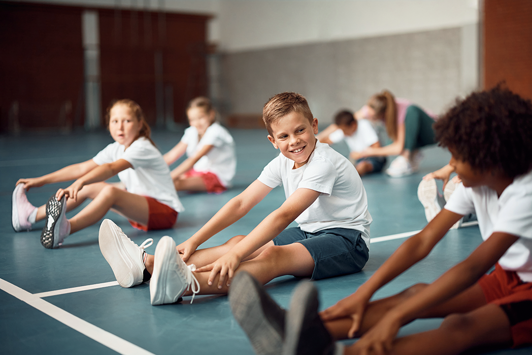 Children, in PE class, sitting in a sports hall stretching their hamstrings by trying to touch their toes.
